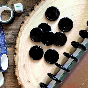 Op de afbeelding: Een close-up van een set zwarte, schijfvormige planneraccessoires. De schijven zijn gerangschikt op een houten oppervlak met een witte dobbelsteen die het nummer "1" laat zien op de achtergrond.