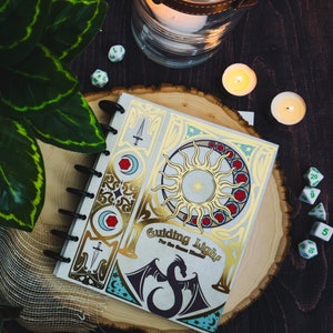 May include: A gold and white journal with a decorative design featuring a sun, moon, and dragon. The journal has a gold foil title that reads "Guiding Light For the Game Master".