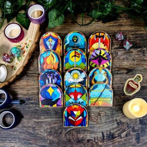 Könnte beinhalten: Ein Set aus 15 bunten Buntglasfenstern mit verschiedenen Symbolen und Designs. Die Fliesen sind in einem Halbkreis auf einer Holzoberfläche angeordnet. Jede Fliese hat ein anderes Design, darunter ein Schwert, eine Krone, ein Herz, ein Stern und eine Sonne.