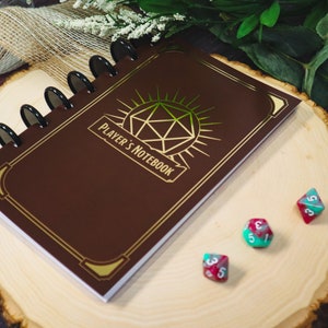 Peut inclure: Un carnet en cuir marron avec une reliure spirale dorée et un motif doré d'un dé à vingt faces avec les mots "Player's Notebook" sur la couverture. Trois dés rouges et verts sont sur une surface en bois.