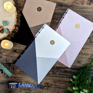 May include: Three different notebooks with geometric designs and gold foil accents. The notebooks have a brown, pink, and gray color scheme. Each notebook has a gold foil die symbol on the cover.