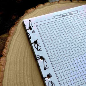 Pode incluir: Um caderno branco quadriculado "Dungeon Map" com encadernação decorativa em metal preto. O caderno está aberto, revelando o padrão de grade. A encadernação apresenta várias formas recortadas em preto. O caderno repousa sobre uma superfície de madeira.