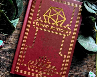 May include: A burgundy notebook with gold accents and a gold die design on the cover. The text "Player's Notebook" and "Adventure Awaits" is printed in gold on the cover.