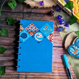 Könnte beinhalten: Ein blaues Spiralnotizbuch mit einem dekorativen geometrischen Design in Orange, Rot und Weiß. Das Notizbuch ist offen und zeigt die schwarze Spiralbindung. Ein türkisfarbener Stift und mehrere Würfel sind ebenfalls sichtbar.