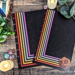 Puede incluir: Dos cuadernos negros con cubiertas rayadas de colores. Los cuadernos tienen un sistema de encuadernación de discos y están abiertos para revelar páginas en blanco. Los cuadernos están sobre una superficie de madera con dados y velas.