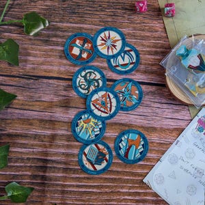 May include: A collection of circular game tokens with blue and white borders, featuring various symbols and text. The tokens are arranged on a wooden surface, alongside dice, a small wooden coaster, and a piece of paper with handwritten notes.