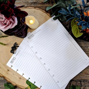 May include: Lined white paper with the word "NOTES" printed at the top, designed for a binder. The paper is on a wooden surface, with a decorative arrangement of flowers and a candle in the background. A black spider decoration is also present.