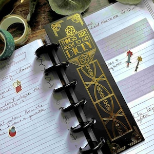May include: A black and gold bookmark with the text "Things are about to get dicey" and a gold design of dice. The bookmark is attached to a black and silver ring binder.