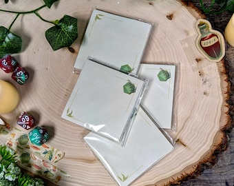 Op de afbeelding: Vier blanco, lege notitiekaartjes met groene bladeren en een groen-wit geometrisch patroon. De kaartjes liggen op een houten oppervlak met een rood-witte dobbelsteen en een klein rood flesje met een wit etiket waarop "4D414" staat.