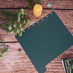 May include: A dark green rectangular paper sheet with a series of circular punch holes along one edge. The paper is positioned near a small yellow LED candle, a cluster of white flowers, two multi-sided dice, and a book titled Tome of Spells.