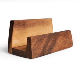 May include: A wooden business card holder with a natural finish. The holder is rectangular with a curved back and a front lip to hold cards. The wood grain is visible, and the holder is set against a white background.