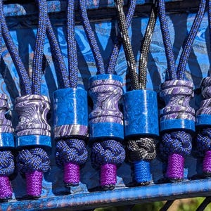 May include: Close-up of several lanyard beads with blue, purple, and silver accents. The beads have a variety of shapes and patterns, with knotted rope and purple tassels. The lanyards are displayed on a blue surface.