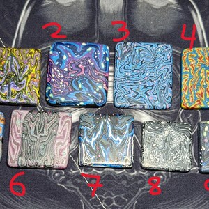 May include: Nine square-shaped pieces of colorful, marbled, and swirled polymer clay. The pieces are arranged in three rows of three. The colors include blue, yellow, red, black, white, and pink.