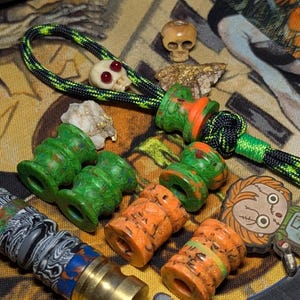 May include: A collection of decorative beads and charms in various colors and designs. Includes green, orange, and black beads, a skull charm with red eyes, and a Chucky-themed charm. The beads are made of different materials and have unique patterns.