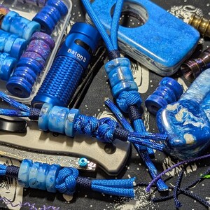 May include: A collection of blue resin beads with marbled patterns, some attached to blue paracord lanyards with knots. A blue Baton 3 flashlight, a blue resin cigar cutter, and a metallic folding knife are visible among the various cylindrical beads.