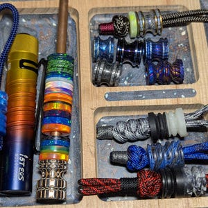 May include: A wooden tray with various colorful beads, a flashlight, a pen, and a metal object with a ring. The beads are made of different materials, including metal, plastic, and wood. The flashlight is black and has a silver ring around it. The pen is brown and has a silver tip. The metal object is silver and has a ring around it.