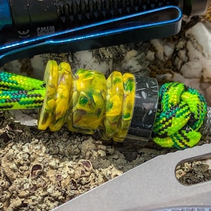 May include: Close-up of a paracord lanyard with a yellow and green marbled bead, a black and gray bead, and a green and black knotted section. A silver knife and a black and blue flashlight are in the background.