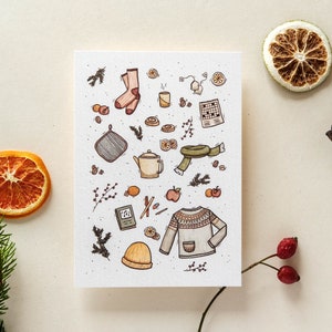 Könnte beinhalten: Eine weiße Karte mit einer bunten Illustration von gemütlichen Winterartikeln, darunter eine Teekanne, ein Pullover, Socken, ein Hut und eine Tasse. Die Illustration ist von kleinen Details wie Beeren, Blättern und Orangen umgeben.