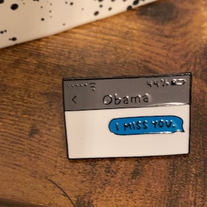 Könnte beinhalten: Eine silberne Emaille-Anstecknadel in Form eines Computerbildschirms mit dem Text "Obama" und einer blauen Sprechblase, die "Ich vermisse dich" sagt.