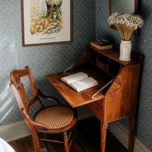 May include: A vintage wooden desk with a drop-down writing surface and a matching chair with a woven seat. A framed illustration hangs on the wall above the desk. A vase of dried flowers sits on the desk. The room has blue floral wallpaper.