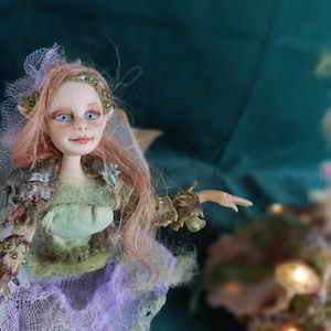 Può includere: Una bambola con lunghi capelli rosa, occhi azzurri luminosi e lentiggini, che indossa un top verde chiaro e una gonna a strati in rete viola. La bambola presenta orecchie a punta, una fascia decorativa con perline verdi e dettagli sulle braccia.