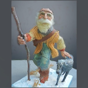 Può includere: Una statuetta fatta a mano di un uomo barbuto con una lunga barba bianca, che indossa un cappello marrone, un maglione giallo e pantaloni blu. Tiene un bastone nella mano destra e cammina con un cane nero al suo fianco. La statuetta è posizionata su uno sfondo innevato.