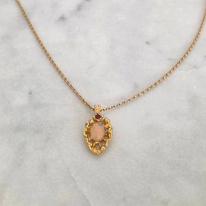 May include: Gold-toned necklace with a pendant featuring a peach-colored stone and a small red accent stone. The pendant has an ornate, textured frame. The necklace is displayed on a white marble surface.