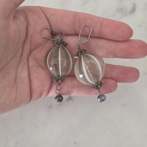 May include: A pair of dangle earrings featuring clear, orb-shaped glass beads with vertical white stripes. The earrings have silver-toned metal accents and a small, dark gray bead at the bottom. The earrings are held in a hand.