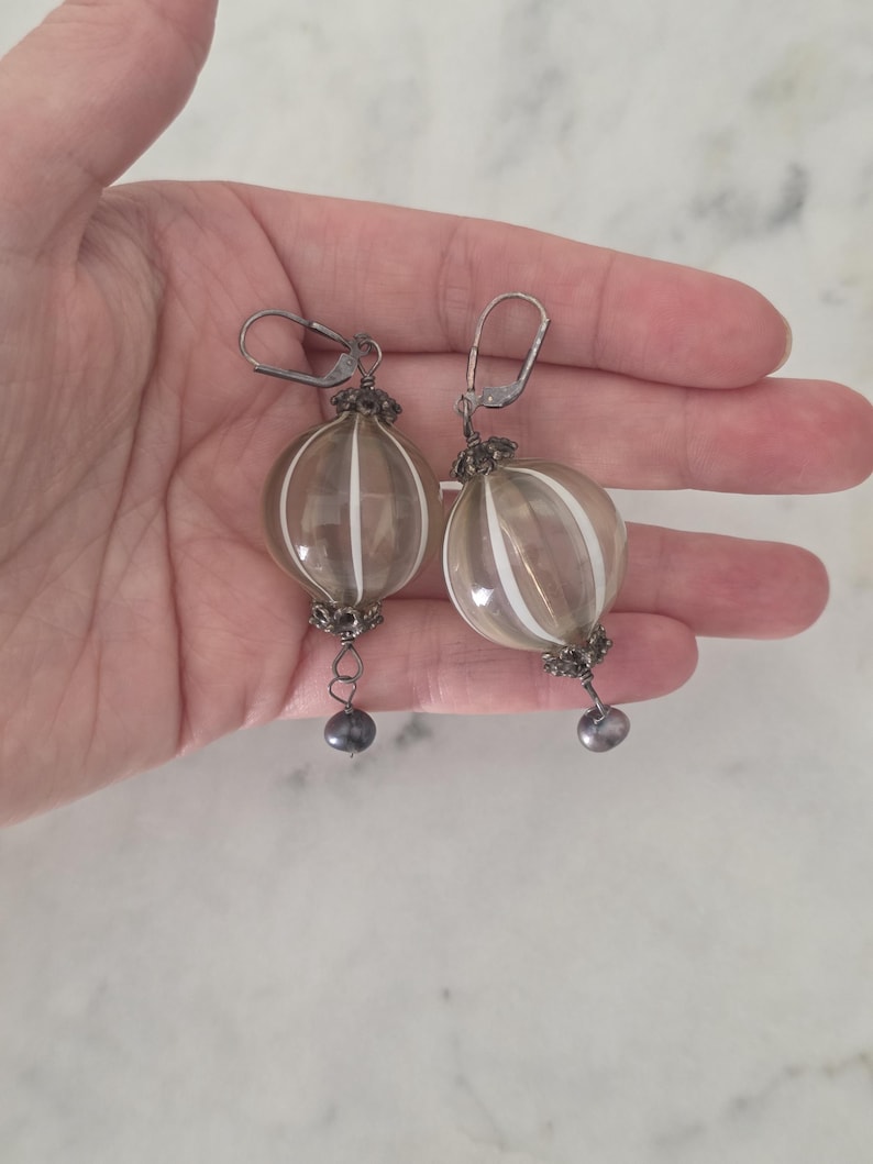 May include: A pair of dangle earrings featuring clear, orb-shaped glass beads with vertical white stripes. The earrings have silver-toned metal accents and a small, dark gray bead at the bottom. The earrings are held in a hand.