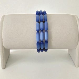 Könnte beinhalten: Ein dreireihiges Armband mit länglichen, oval geformten Perlen in verschiedenen Blautönen. Die Perlen sind aneinandergereiht und erzeugen einen geschichteten Effekt. Das Armband wird auf einem weißen Ständer präsentiert, um sein Design und seine Farbvariationen hervorzuheben.