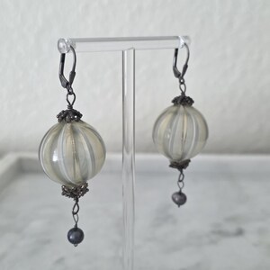May include: A pair of dangle earrings featuring round, ribbed, light-colored glass orbs with silver-tone metal accents. Each earring has a dark gray bead at the bottom and a lever-back closure. The earrings are displayed on a clear stand.