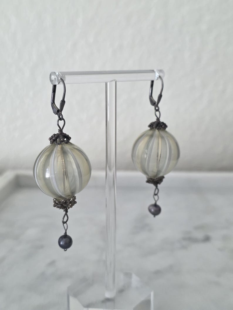 May include: A pair of dangle earrings featuring round, ribbed, light-colored glass orbs with silver-tone metal accents. Each earring has a dark gray bead at the bottom and a lever-back closure. The earrings are displayed on a clear stand.