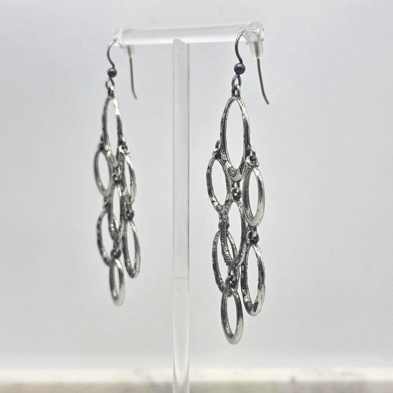 May include: Silver-toned chandelier earrings with multiple oval-shaped links. The earrings have a textured finish and dangle from a simple hook. The design is symmetrical, with each earring featuring a series of connected ovals.