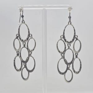 May include: Silver-toned chandelier earrings with an oval design. Each earring features multiple oval shapes connected in a cascading arrangement. The metal has a textured appearance, and the earrings hang from a simple hook.