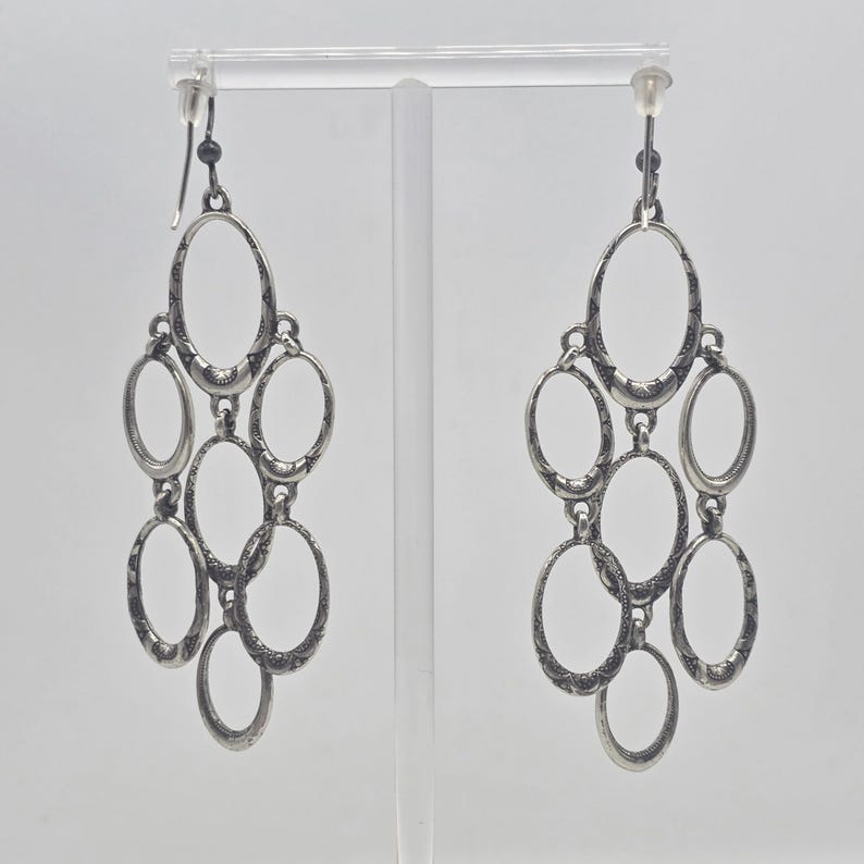 May include: Silver-toned chandelier earrings with an oval design. Each earring features multiple oval shapes connected in a cascading arrangement. The metal has a textured appearance, and the earrings hang from a simple hook.