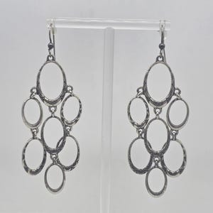May include: A pair of silver-toned chandelier earrings. Each earring features multiple oval-shaped frames connected in a cascading design. The metal has an antique finish, and the earrings hang from a simple hook.