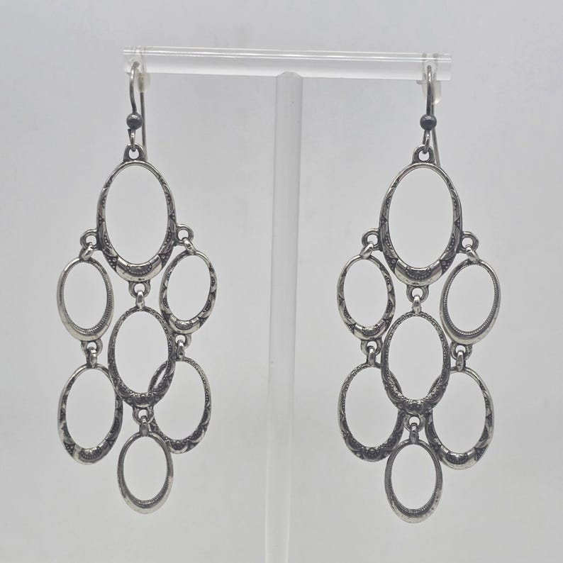 May include: A pair of silver-toned chandelier earrings. Each earring features multiple oval-shaped frames connected in a cascading design. The metal has an antique finish, and the earrings hang from a simple hook.
