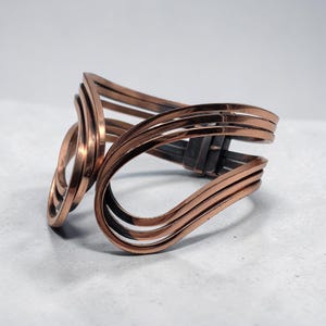 May include: A copper-colored cuff bracelet with a unique, open design. The bracelet features multiple curved bands that create an open, flowing shape. The metal has a polished finish, reflecting light and adding to its visual appeal. The bracelet is a statement piece.