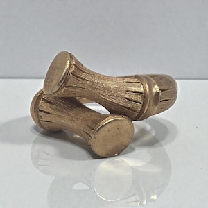May include: A gold-colored ring with a unique design. The ring features two cylindrical elements with a textured, wood-like appearance, connected by a band. The ring is displayed on a reflective surface, highlighting its metallic sheen.