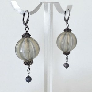 May include: A pair of dangle earrings featuring a round, ribbed, light-colored glass orb with a dark metal filigree cap and a small, dark bead at the bottom. The earrings are displayed on a clear stand against a white background.