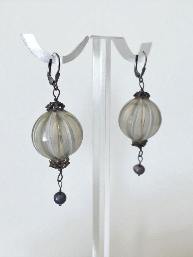 May include: A pair of dangle earrings featuring a round, ribbed, light-colored glass orb with a dark metal filigree cap and a small, dark bead at the bottom. The earrings are displayed on a clear stand against a white background.