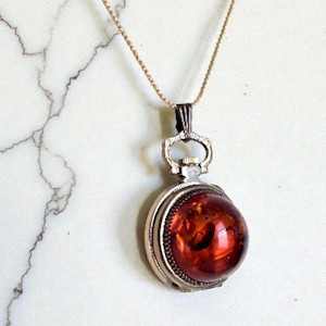 May include: A vintage pendant necklace featuring a round, amber-colored stone encased in a silver-toned metal setting. The pendant hangs from a delicate gold-colored chain. The stone has a warm, reddish-orange hue, and the setting has intricate detailing.
