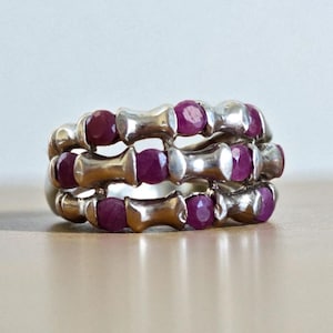 Puede incluir: Tres anillos de plata apilados, cada uno con pequeñas gemas redondas de color rojo intenso. Los anillos tienen un diseño único con segmentos plateados entre las gemas, creando un aspecto distintivo. Los anillos se exhiben sobre una superficie clara.