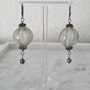 May include: A pair of handcrafted earrings featuring round, striped orbs in shades of gray and white. The earrings have dark metal accents and a small, dark gray pearl at the bottom. They are displayed on a clear acrylic stand.