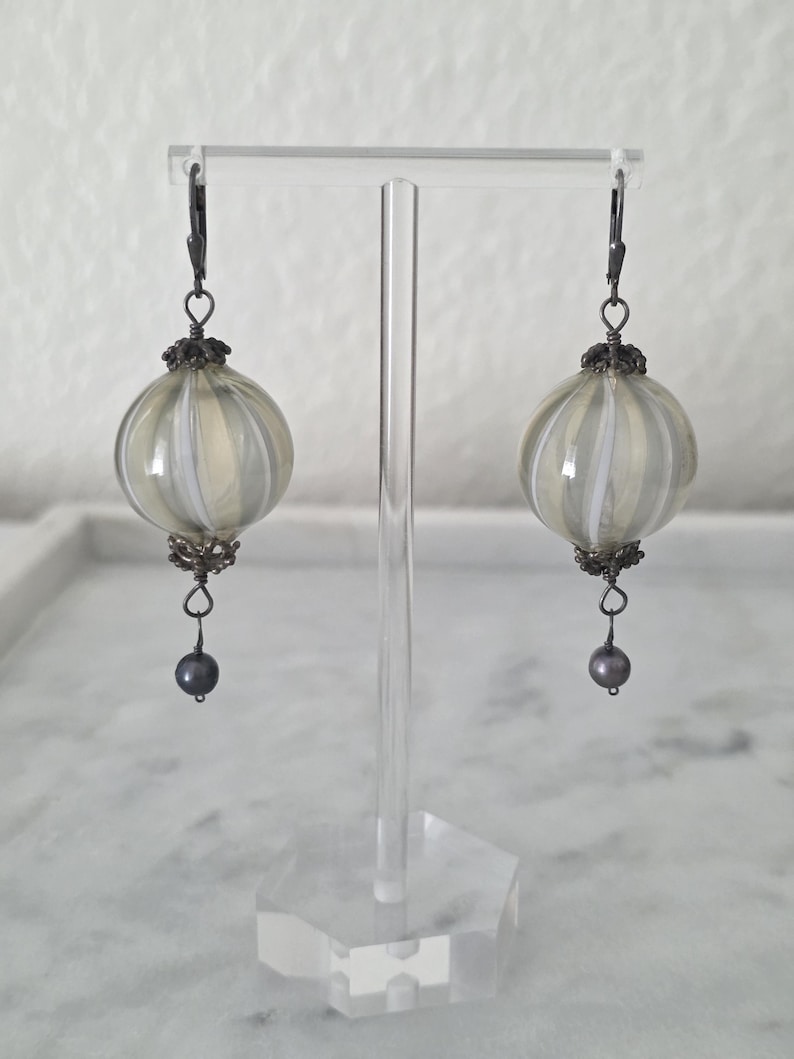 May include: A pair of handcrafted earrings featuring round, striped orbs in shades of gray and white. The earrings have dark metal accents and a small, dark gray pearl at the bottom. They are displayed on a clear acrylic stand.
