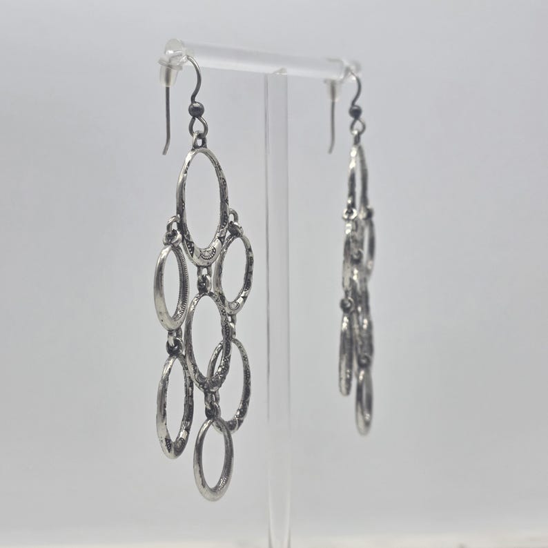 May include: Silver-toned chandelier earrings with multiple oval-shaped links. The earrings have a textured finish and dangle from a simple hook. The design is intricate and detailed, suitable for a formal or casual look.
