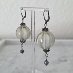 May include: A pair of earrings with a vintage aesthetic. Each earring features a round, ribbed, cream-colored glass orb with white stripes. The earrings have dark metal accents and a small, dark gray bead dangling from the bottom. They are displayed on a clear stand.