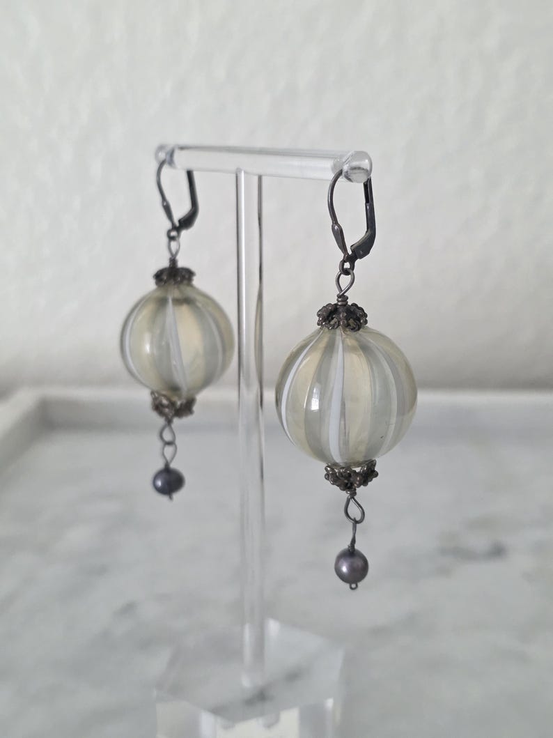 May include: A pair of earrings with a vintage aesthetic. Each earring features a round, ribbed, cream-colored glass orb with white stripes. The earrings have dark metal accents and a small, dark gray bead dangling from the bottom. They are displayed on a clear stand.