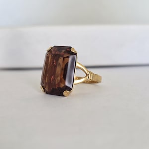 Puede incluir: Anillo de oro con una gran gema rectangular de color marrón. La gema está engastada en una banda de oro. El anillo se exhibe sobre una superficie blanca, mostrando su color y forma.