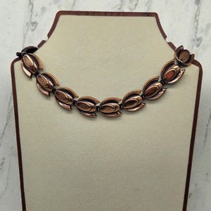 Op de afbeelding: Een koperkleurige ketting met een bloemmotief, tentoongesteld op een mannequin. De ketting heeft een reeks gestileerde bloemvormige schakels die zijn verbonden door kleine, donkergekleurde connectoren. Het algehele ontwerp is symmetrisch en vintage-geïnspireerd.